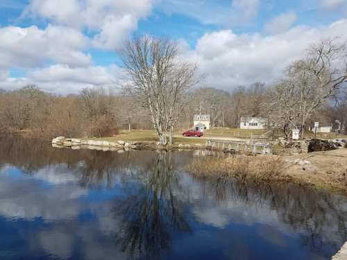 A serene landscape featuring a calm pond reflecting trees and clouds, with two houses in the background.