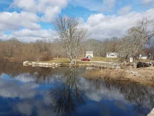 A serene landscape featuring a calm pond reflecting trees and clouds, with two houses in the background.