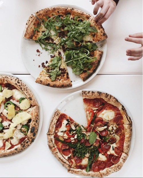 Three pizzas on white plates: one with pepperoni and veggies, one with pineapple and greens, and a third with arugula.