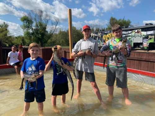 Four children stand in shallow water, each holding a small alligator, with trees and a food truck in the background.