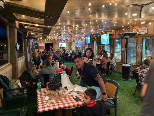 A lively restaurant with string lights, families dining, and a man smiling at the camera, creating a warm atmosphere.