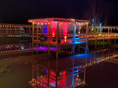 A brightly lit wooden pavilion at night, reflecting in water, surrounded by colorful lights and decorations.