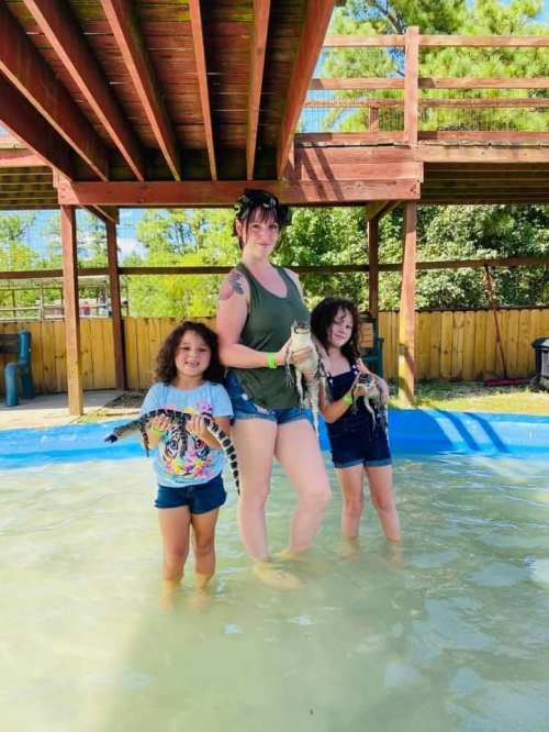 A woman and two girls stand in shallow water, each holding a small alligator, with a wooden deck in the background.