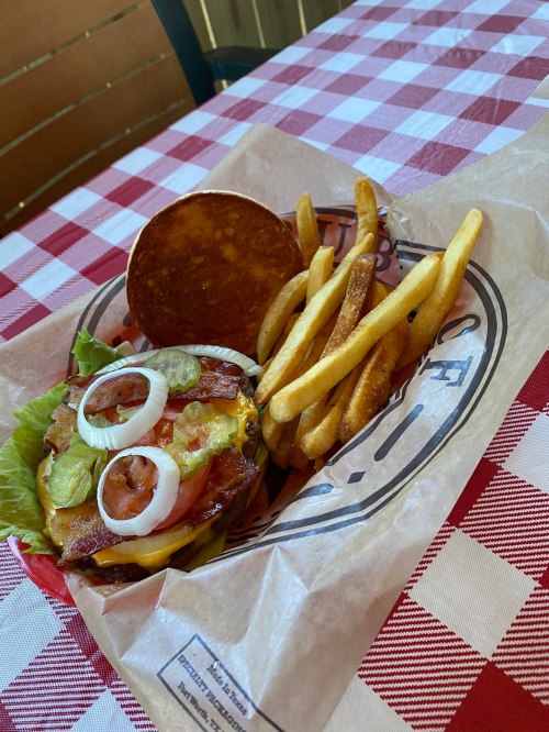 A burger with lettuce, bacon, cheese, and onions, served with a side of golden fries on a checkered tablecloth.