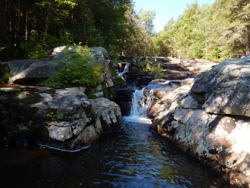 A serene waterfall cascades over rocky terrain, surrounded by lush greenery and sunlight filtering through the trees.