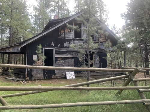 Abandoned, weathered house surrounded by trees, with a wooden fence in front and a target board on the ground.