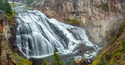 A cascading waterfall flows over rocky cliffs, surrounded by lush greenery and mist in a serene natural landscape.
