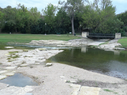 A serene park scene featuring a shallow stream, rocky banks, and a small bridge surrounded by greenery.
