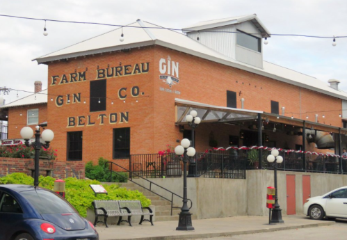 Historic brick building with "Farm Bureau Gin Co." sign, outdoor seating, and decorative lights in Belton.