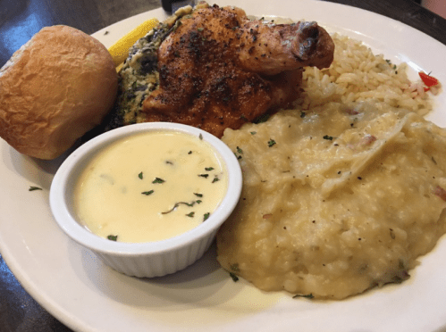 A plate featuring seasoned chicken, rice, mashed potatoes, a roll, and a small bowl of creamy sauce.