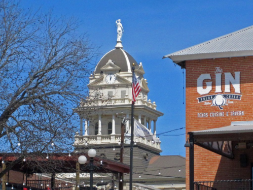 Historic clock tower with a statue on top, blue sky, and a restaurant sign reading "The Gin" in the foreground.