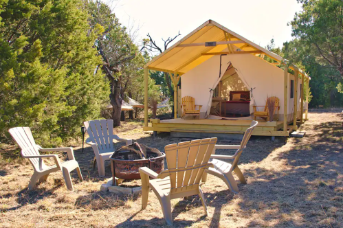 A cozy glamping tent with a wooden deck, surrounded by trees and a fire pit with chairs in a serene outdoor setting.