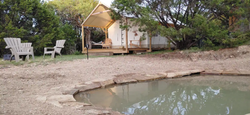 A cozy cabin with a porch, surrounded by trees, next to a small pond and outdoor seating area.