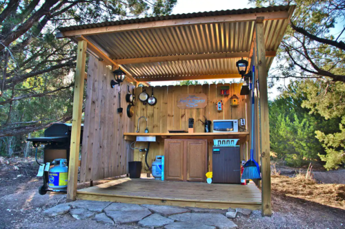 A rustic outdoor kitchen with wooden walls, a metal roof, and various appliances, set in a natural landscape.