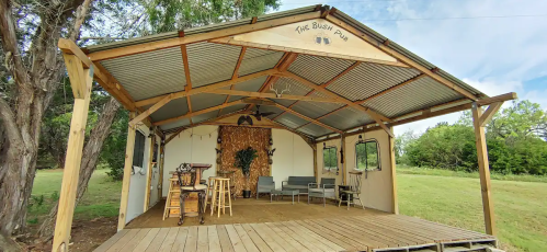 A rustic outdoor structure with a wooden deck, seating area, and decorative wall, surrounded by greenery.