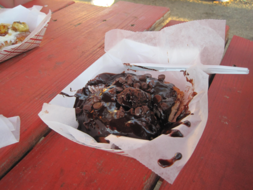 A dessert in a paper tray, featuring chocolate sauce, chocolate chips, and a brownie on a red picnic table.