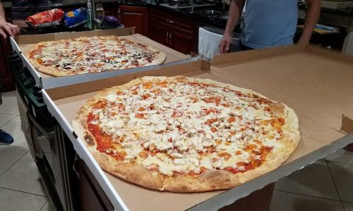 Two large pizzas on a kitchen counter, one topped with chicken and the other with various toppings, surrounded by people.