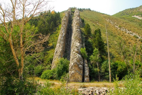 Two tall rock formations rise from a hillside, surrounded by greenery and trees under a clear sky.