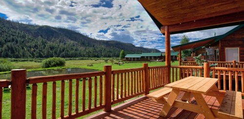 A wooden deck with a picnic table overlooks a lush green valley and mountains under a partly cloudy sky.