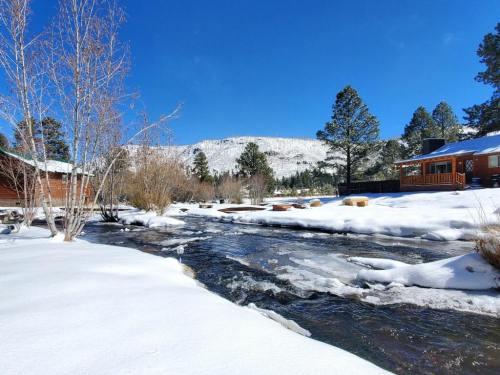 A snowy landscape with a flowing river, surrounded by trees and mountains under a clear blue sky.