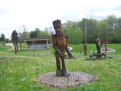 A wooden sculpture of a figure stands in a grassy park, surrounded by trees and a small pavilion in the background.