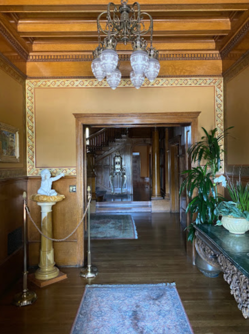 Elegant hallway with wooden paneling, chandelier, and decorative plants, leading to a staircase in the background.