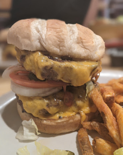 A towering cheeseburger with two patties, melted cheese, lettuce, tomato, and onion, served with a side of fries.