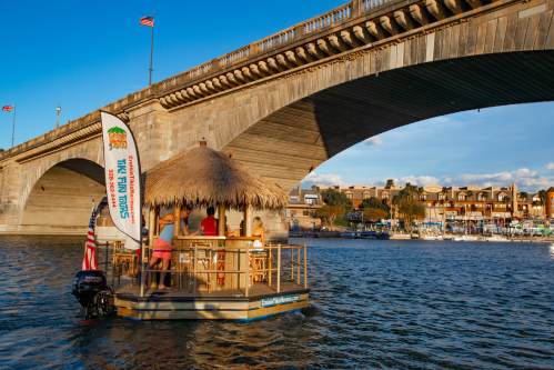 A small boat with a thatched roof floats under a bridge, with people enjoying the view and flags waving in the breeze.