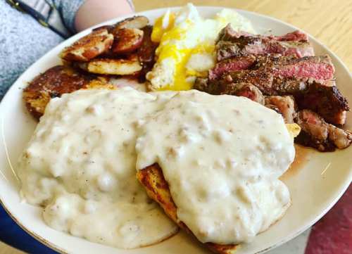 A plate of breakfast featuring steak, eggs, potatoes, and biscuits smothered in creamy gravy.