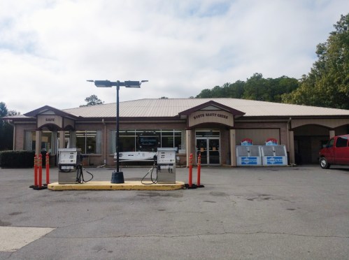 A gas station with fuel pumps in front of a building, featuring a cafe and convenience store. Trees in the background.
