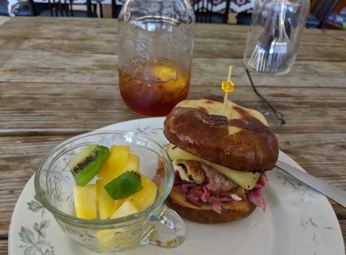 A sandwich with meat and pineapple on a plate, accompanied by a fruit cup and a glass of iced tea.