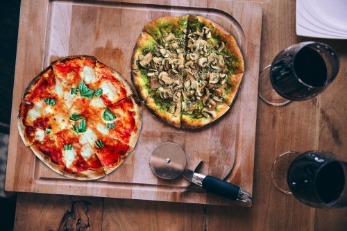 A wooden platter with two pizzas: one with tomato sauce and cheese, the other topped with mushrooms and green sauce, alongside two glasses of red wine.