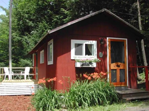 A small red cabin surrounded by greenery, featuring a wooden door, flower boxes, and a deck with chairs.