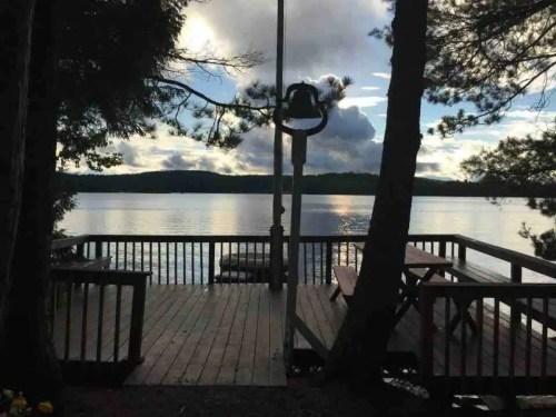 A serene lakeside view from a wooden deck, surrounded by trees and reflecting clouds on the water's surface.