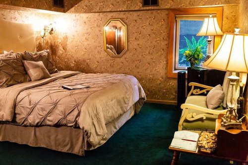 Cozy bedroom with a large bed, decorative wallpaper, a window, and a small seating area with lamps.