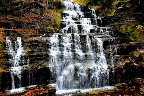 A cascading waterfall flows over rocky cliffs, surrounded by autumn foliage in vibrant colors.