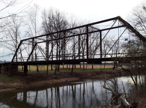 An old, rusted metal bridge spans a calm river, surrounded by bare trees and open fields in the background.