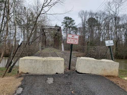 A closed bridge with "No Trespassing" signs and concrete barriers, surrounded by trees and a river.
