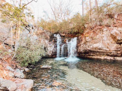 A serene waterfall cascades over rocks into a clear pool, surrounded by trees and autumn foliage.