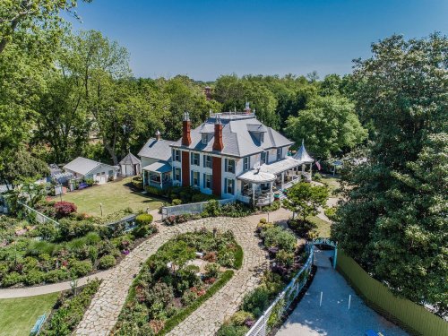 Aerial view of a large historic house surrounded by lush gardens and trees on a clear sunny day.