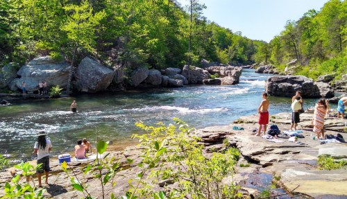 People enjoying a sunny day by a river, surrounded by rocks and greenery, with some swimming and others relaxing.