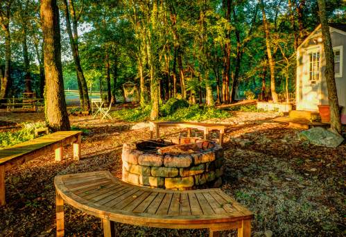 A cozy outdoor space with a stone fire pit, wooden benches, and trees, near a serene lake in the background.