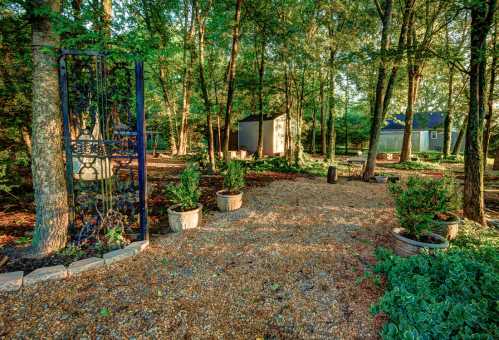 A serene garden scene with trees, gravel paths, potted plants, and small structures in a peaceful, natural setting.