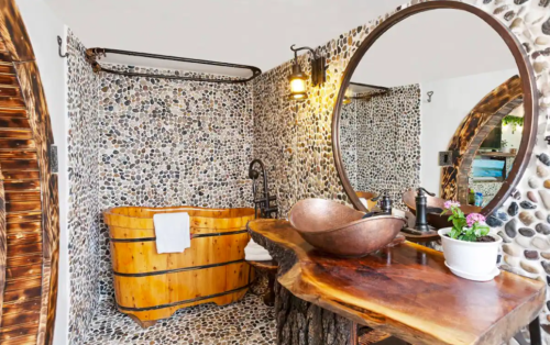 A rustic bathroom featuring a wooden bathtub, stone walls, a round mirror, and a wooden sink with a plant.