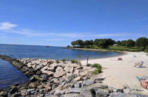 A sunny beach scene with a rocky shoreline, calm water, and people relaxing on the sand under a clear blue sky.