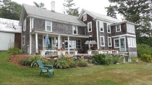 A charming two-story house with a shingled exterior, surrounded by a garden and lawn furniture.