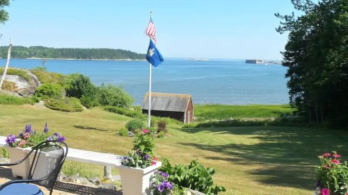 A serene coastal view featuring a flag, a red shed, and lush green grass by the water under a clear blue sky.