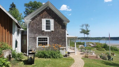 A charming wooden house with a garden, overlooking a serene waterfront and blue sky.