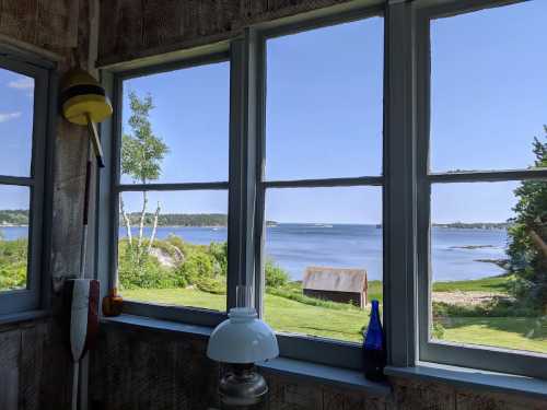 View from a window showing a serene coastal landscape with water, greenery, and a small shed in the distance.