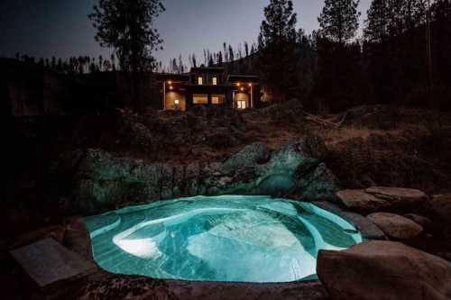 A serene outdoor hot tub surrounded by rocks, with a cozy house illuminated in the background at dusk.